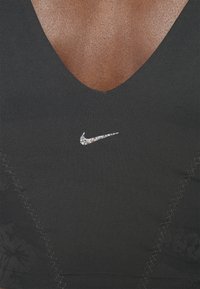 Reggiseno sportivo nero con scollo a V profondo, accentuato da una texture floreale e un logo Nike argento brillante al centro. Tessuto liscio con dettagli di cucitura.