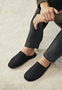 Chaussons noirs à enfiler avec une texture côtelée et un design arrondi, dotés d'un intérieur doux et d'une semelle en caoutchouc plate, posés sur un tapis clair.