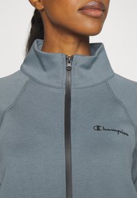 Ljusblå zip-jacka med hög krage, svart dragkedja och logotyp. Slät textur med kontrasterande sömmar och figurnära design.