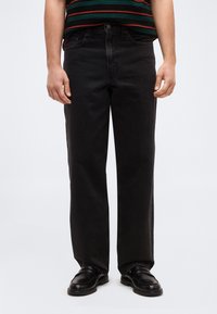 Άνδρας με μαύρο τζιν  ίσιας γραμμής, μαύρα δερμάτινα μοκασίνια και ριγέ πουκάμισο, στέκεται με τα χέρια χαλαρωμένα στα πλευρά.