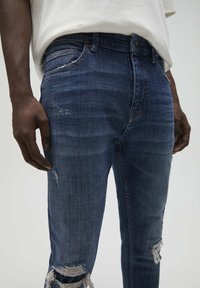Blue denim jeans with distressed detailing, a straight cut, five pockets, and a mid-rise waist. The fabric shows subtle fading and a soft texture.