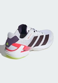 Chaussure de sport blanche avec des rayures noires, semelle néon verte et accents rouges et violets, présentée de côté sur un fond clair.