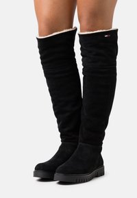 Stivali alti fino alla coscia in suede nero con una morbida fodera bianca, suola piatta nera e dettagli testurizzati. Design semplice senza hardware visibile.