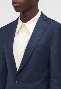 Homme portant un blazer bleu foncé texturé par-dessus une chemise crème boutonnée avec un large col, présenté des épaules au menton sur un fond uni.