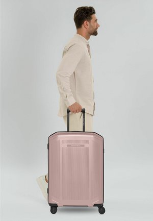 Mann in beiger Kleidung zieht einen großen hartschaligen Koffer in Roségold mit Teleskopgriff vor weißem Hintergrund.