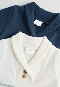 Dve srajci z ovratnikom: ena temno modra in ena kremne barve, z rebrastimi ovratniki, gumbi in vidnimi oznakami blagovne znamke.