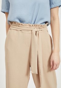 Beżowe spodnie z wysokim stanem, z falbaną w pasie i wiązanym paskiem. Materiał jest lekki i ma gładką fakturę.