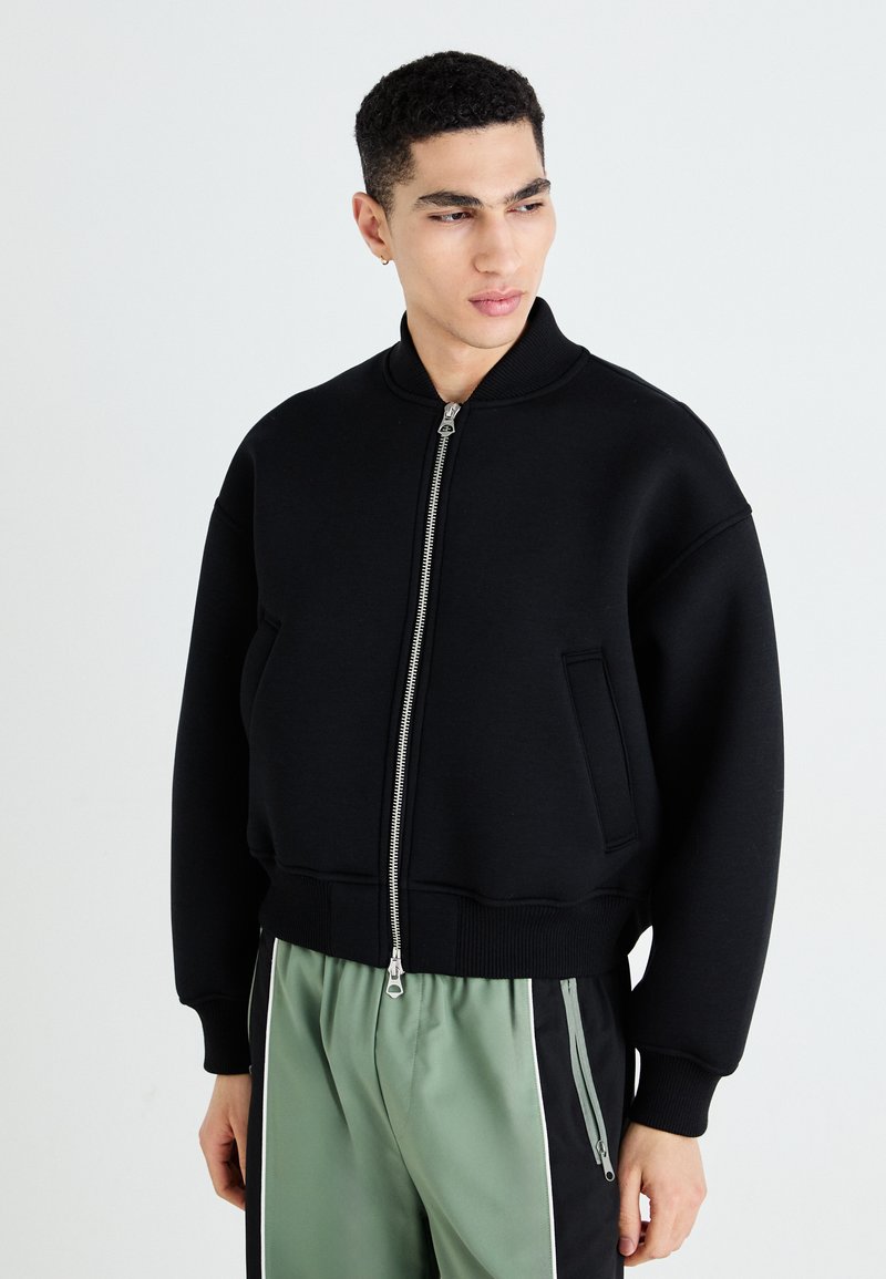 Veste bomber noire avec un col haut, fermeture éclair sur le devant, deux poches latérales et poignets côtelés. Le tissu semble lisse et épais.