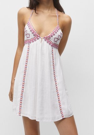 Vrouw draagt een witte mouwloze jurk met roze en paarse geborduurde patronen en dunne schouderbandjes.