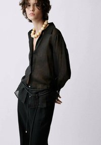 Black sheer long-sleeve shirt with an open collar, paired with black pants and a textured rope belt. Features a large beige bead necklace.