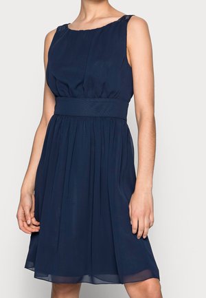 Femme portant une robe bleu marine sans manches, longueur genou, avec une taille ajustée et une jupe plissée, debout devant un fond clair.