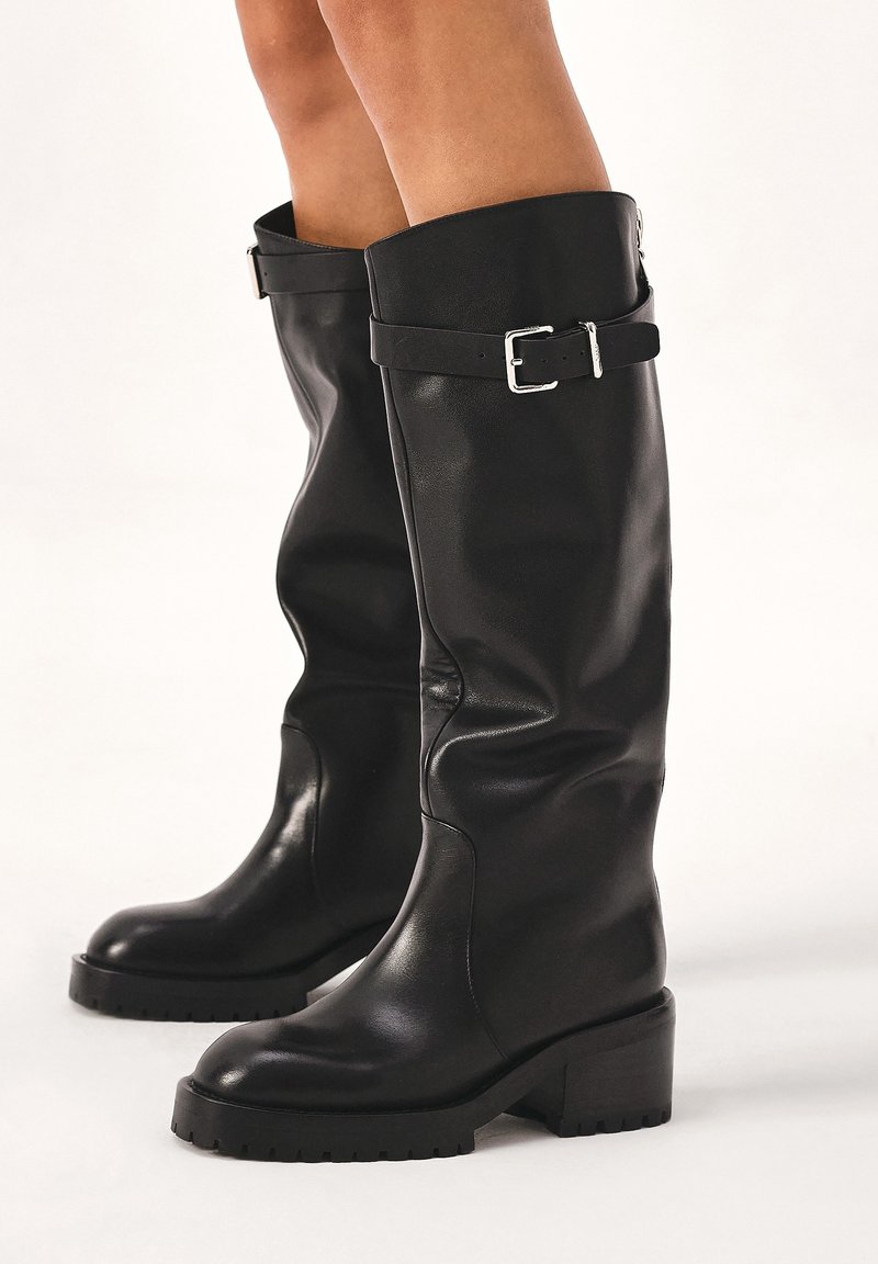 Bottes noires en cuir jusqu'aux genoux avec un talon épais, ornées d'une sangle et d'une boucle au sommet pour un design supplémentaire. Texture lisse et semelle durable.