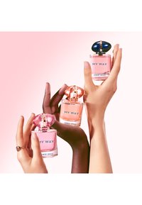 Drei unterschiedliche Hände halten pinke "My Way" Giorgio Armani Parfümflaschen vor einem sanften rosa Hintergrund.