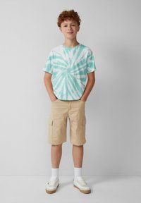 Koszulka z krótkim rękawem w barwy tie-dye w miętowym zielonym i białym kolorze, zestawiona z beżowymi szortami typu cargo. Białe trampki z jasnobrązowymi podeszwami oraz białe skarpetki.