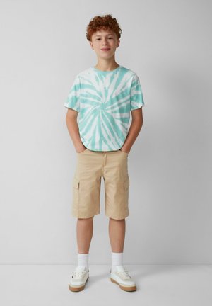 T-shirt à manches courtes en tie-dye vert menthe et blanc, associé à un short cargo beige. Baskets blanches avec des semelles marron clair et chaussettes blanches.