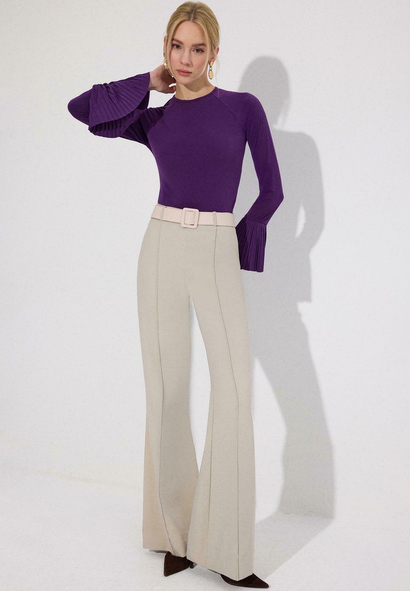 Γυναίκα φορώντας μωβ μπλούζα με μακριά μανίκια και πιέτες στα μανίκια, μπεζ παντελόνι με φαρδιά πόδια και ψηλή μέση, και καφέ αιχμηρά παπούτσια, στέκεται μπροστά σε λευκό τοίχο.