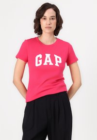 Lyserød bomulds t-shirt med et stort hvidt "GAP" logo på forsiden, rund halsudskæring og korte ærmer, parret med sorte bukser.