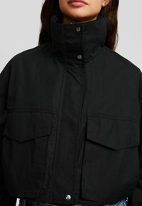 Giacca corta nera realizzata in tessuto resistente, con collo alto, bottoni a pressione e due tasche frontali con patta.