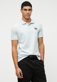 Homem vestindo uma camisa polo azul-claro com um logótipo de óculos de sol pretos no peito, emparelhada com calças pretas e cinto preto, posando contra um fundo branco.