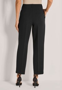 Vrouw in zwarte, op maat gemaakte enkellange broek en zwarte hoge hakken, staande met één hand in de zak.