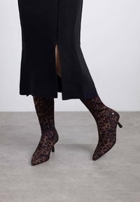 Botas de tacón alto y punta afilada en un tejido transparente con un estampado de leopardo en marrón oscuro y negro; combinadas con un vestido de suéter negro hasta la rodilla.