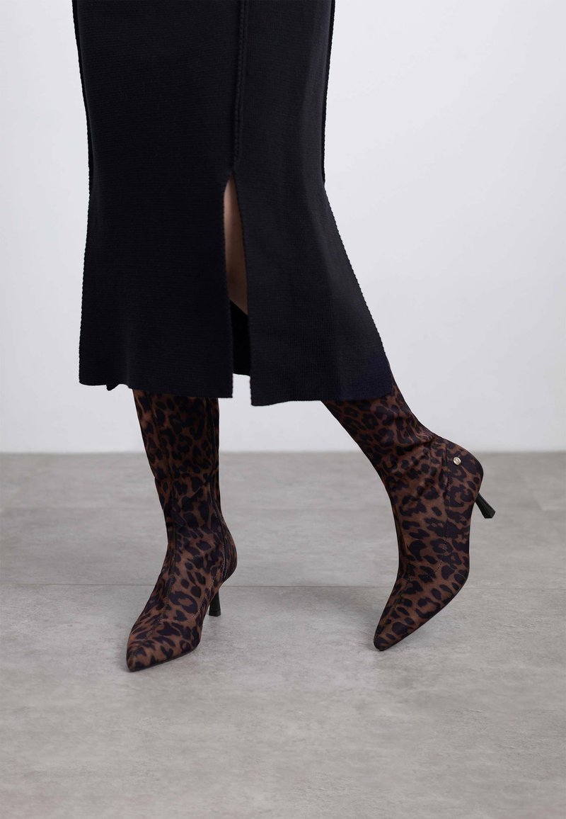 Botas de tacón alto y punta afilada en un tejido transparente con un estampado de leopardo en marrón oscuro y negro; combinadas con un vestido de suéter negro hasta la rodilla.