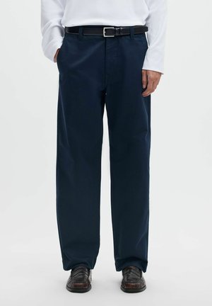 Homme portant un pantalon bleu marine foncé, une ceinture noire, une chemise blanche à manches longues et des chaussures habillées marron foncé, debout avec une main dans la poche.