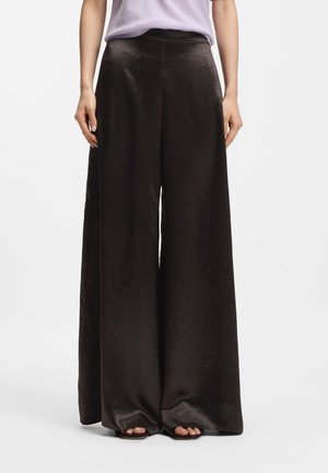 Femme portant un pantalon large en satin noir taille haute associé à un haut violet clair rentré à l'intérieur et des sandales ouvertes.