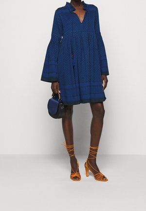 Robe à motif bleu avec des manches cloche et un décolleté à volants, assortie à des talons orange à lanières et un petit sac à main noir.