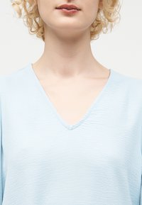 Κοντινό πλάνο ενός ατόμου με ανοιχτόχρωμα σγουρά μαλλιά που φοράει ένα γαλάζιο υφασμάτινο μπλουζάκι με V λαιμόκοψη, με εμφανή τον λαιμό και το πάνω μέρος του στήθους.