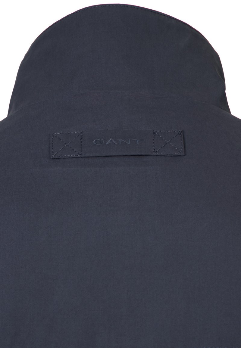 Tessuto blu navy con una targhetta rettangolare cucita e il nome del marchio "GANT" in rilievo al centro, vicino alla parte superiore.