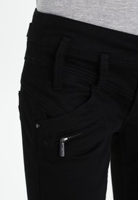Jeans de mezclilla negra con un diseño ajustado, múltiples bolsillos y un detalle de bolsillo con cremallera en el lateral. Textura suave con un acabado oscuro.