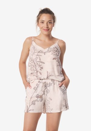 Kobieta stojąca z rękami w kieszeniach, ubrana w jasnoróżowy kwiecisty top na ramiączkach i pasujące szorty, uśmiechająca się na białym tle.