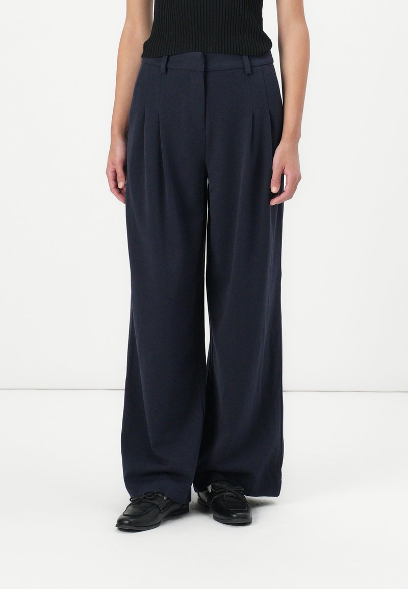 Navy wide-leg trousers with pleats, crafted from a soft fabric. High waist design, side pockets, and a smooth texture. Paired with black shoes.