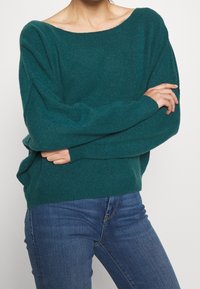 Türkiser Strickpullover mit weitem Ausschnitt, überschnittenen Schultern und gerippten Bündchen. Getragen über blauen Jeans, die eine lässige Passform und weiche Textur zeigen.