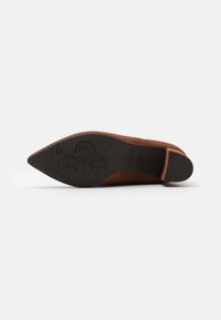 Chaussure beige en daim à bout pointu avec une semelle en caoutchouc noir. Présente des détails de logo embossé sur la semelle et un design épuré et minimaliste.