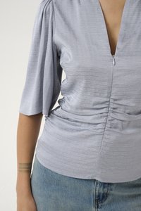 Blusa grigia chiara con un motivo texture, scollatura a V profonda, maniche corte a palloncino e dettaglio arricciato in vita, abbinata a jeans blu.