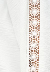 Λευκό ύφασμα με υφή στην επιφάνεια και κάθετη λωρίδα περίτεχνου κυκλικού δαντελωτού μοτίβου με κοψίματα που αποκαλύπτει καφέ φόντο.