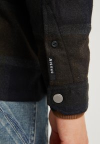 Rukáv kárované vlněné košile s černými a stříbrnými knoflíky a štítkem značky &quot;Chasin&quot;, nošený přes hnědý svetr a modré džíny.