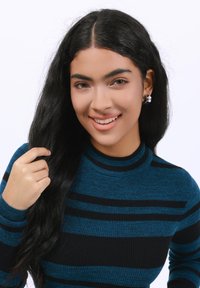 Pull rayé bleu et noir en ribbed avec un col haut. Le modèle a de longs cheveux noirs et porte des boucles d'oreilles rondes en argent.