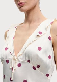 Gros plan sur une robe blanche de femme à pois roses, avec un col en V profond, un col à volants et un détail noué à l’avant.