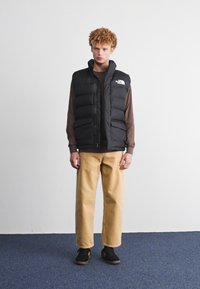 Gilet noir rembourré avec un design matelassé vertical, porté sur une chemise à manches longues marron, associé à un pantalon beige clair et des chaussures noires.