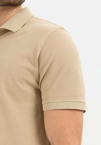 Beżowa koszulka polo z teksturowanej tkaniny, z kołnierzykiem i krótkimi rękawami. Dyskretny logo marki wytłoczone na tkaninie.