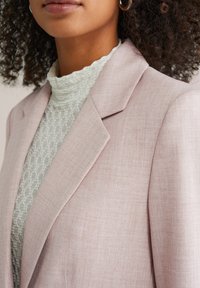 Šviesiai rožinis švarkas su tekstūruotu paviršiumi, turintis įpjautą atlapą ir struktūruotus pečius, dėvimas virš baltos, raštuotos aukšto kaklo palaidinės.