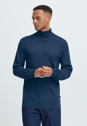 Suéter de cuello alto azul marino en punto acanalado, de mangas largas y con dobladillo recto, combinado con pantalones de denim oscuro. Diseño simple y moderno.