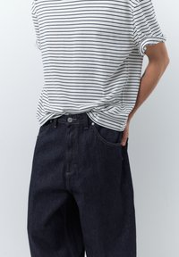 Persona che indossa una maglietta a maniche corte a righe bianche e blu navy infilata in un paio di jeans a gamba larga, a vita alta, di colore blu scuro, con una mano in tasca.