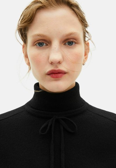 Suéter negro de punto con cuello alto y acento de cordón en el cuello, con ribete acanalado, que presenta una textura suave y un diseño ajustado.