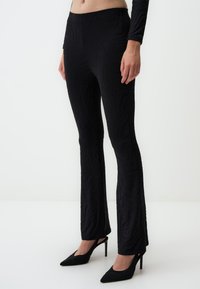 Zwarte flare-broek met een golvend textuurpatroon. Gemaakt van rekbaar materiaal, met een hoge tailleband en gecombineerd met zwarte hakken.