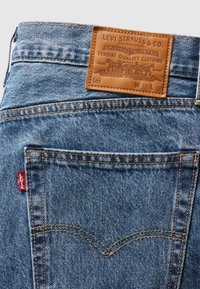 Närbild av blå denim Levi's jeans bakficka med orange sömnad, röd Levi's-lapp och brun lädermärkesetikett på midjan.