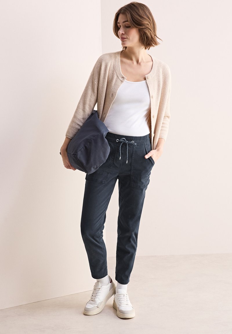Beige stickad kavaj, vit tanktop, mörka byxor med elastisk midja och fickor, samt vita sneakers med grå accenter.